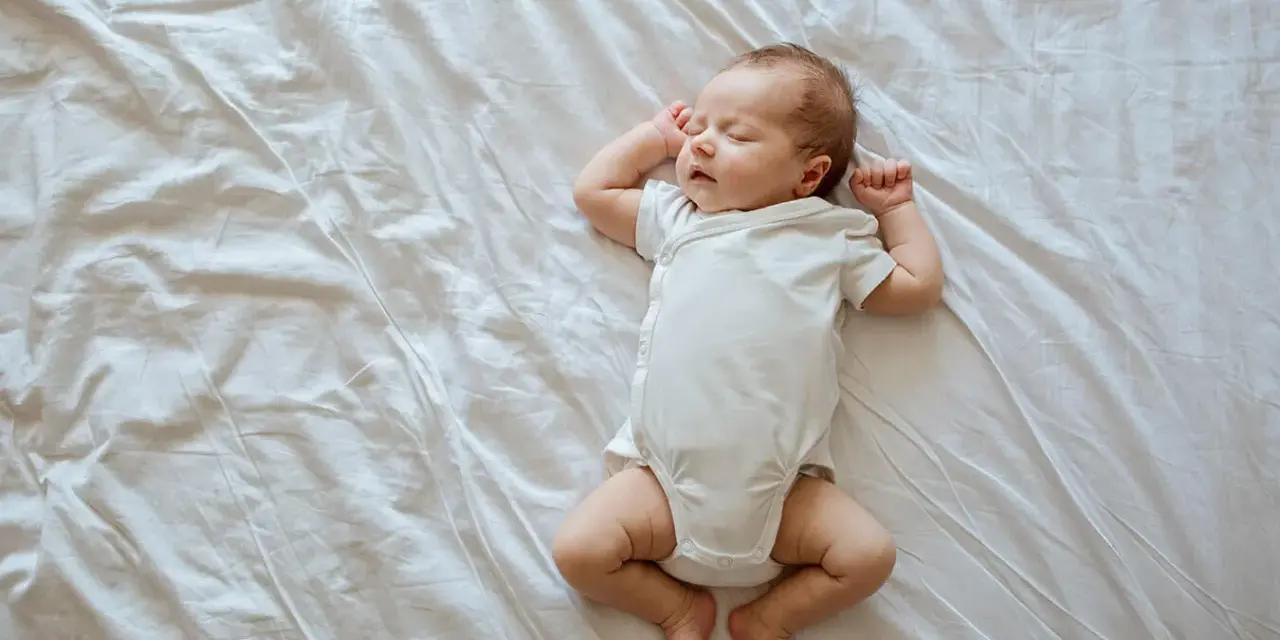 Śpiący, 3 tygodniowy noworodek w białym body leży na pogniecionej białej pościeli.