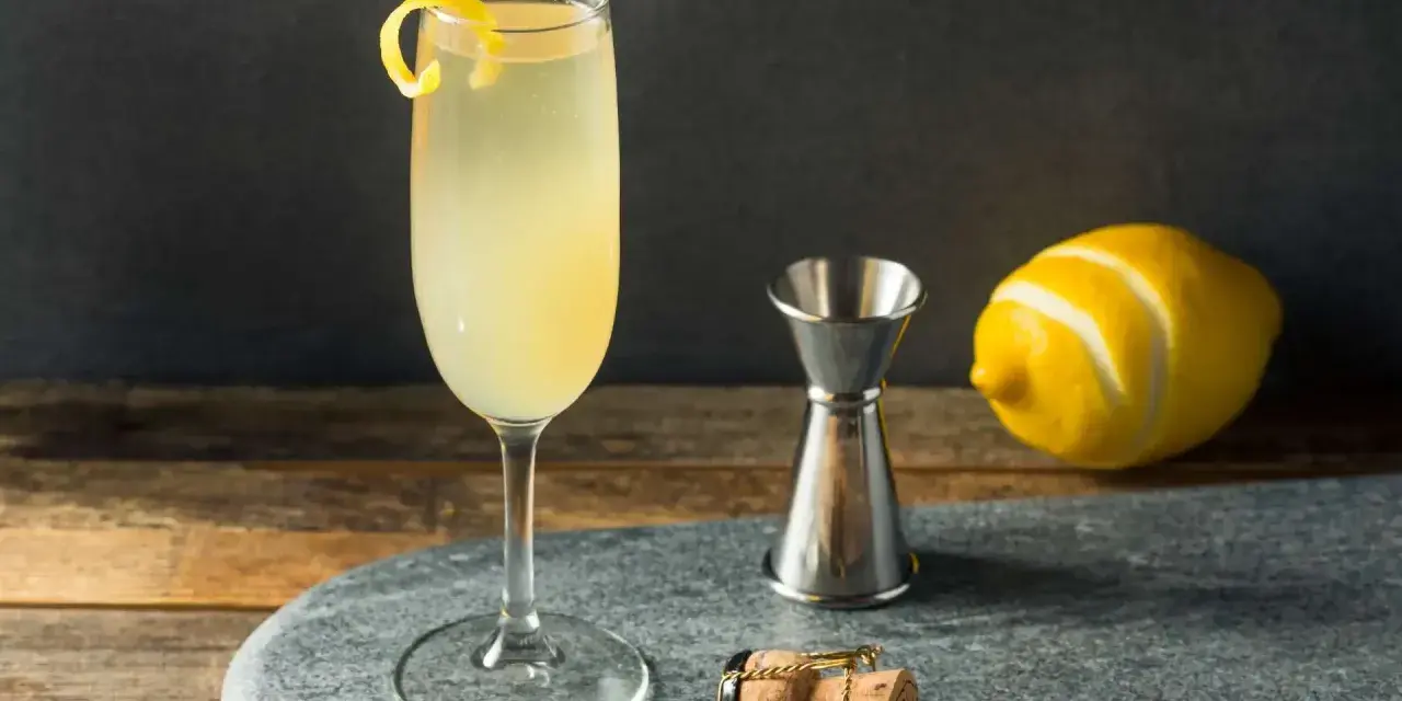 Elegancki drink z szampanem, ozdobiony skórką cytryny, obok kieliszka stoi miarka barmańska, cytryna i korek od szampana.
