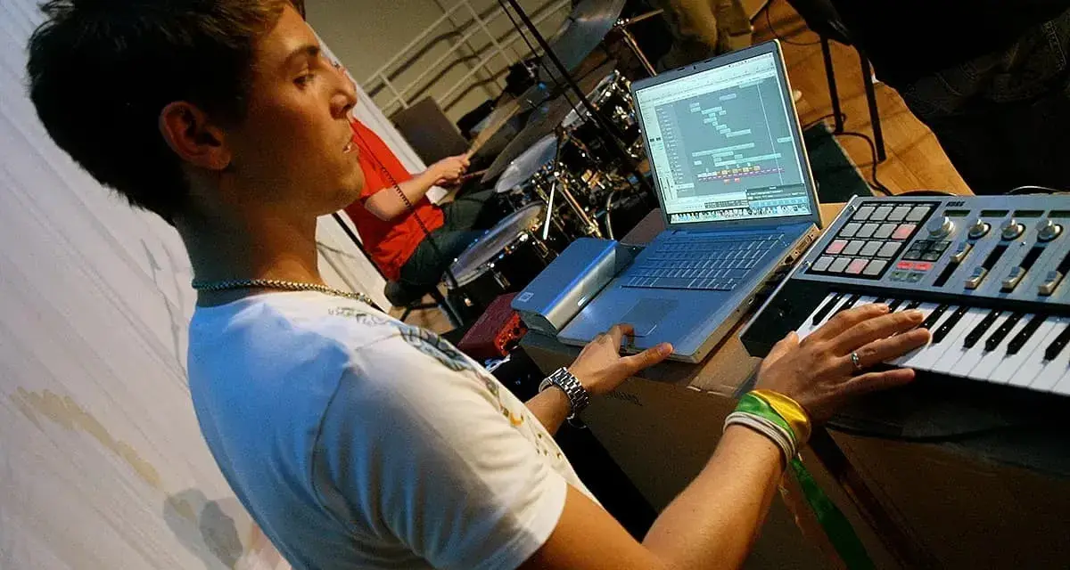 Junger Mann am Laptop und Keyboard, der Musik produziert. Ein Schlagzeuger im Hintergrund. Perfekt für ein Musikproduktion Studium.