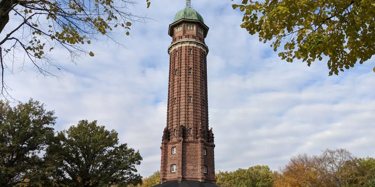 Der Aussichtsturm im Volkspark Jungfernheide ragt mit seinem roten Backstein und grünen Kuppeldach in den Himmel. Herbstlaub umrahmt die Szene.