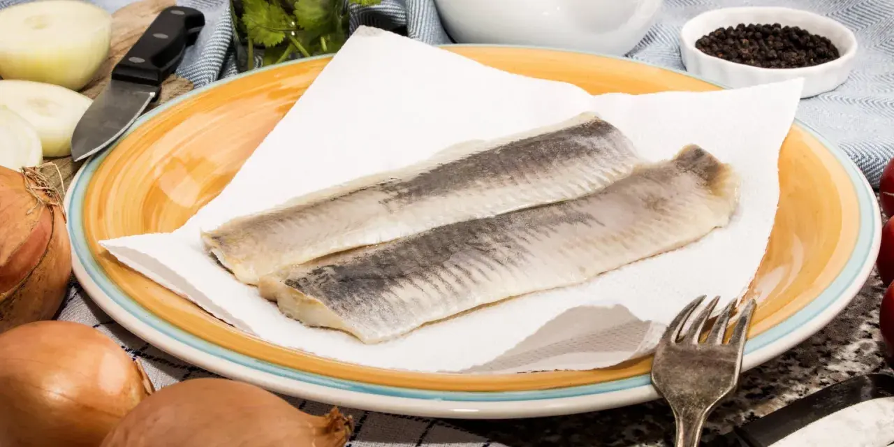 Zwei Matjesfilets auf Küchenpapier, bereit für eine gesunde Mahlzeit. Zwiebeln und Pfefferkörner deuten auf die Zubereitung hin.