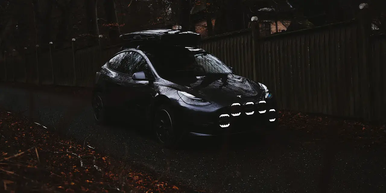 Mörk Tesla med takbox och extraljusstag redo för äventyr längs en höstfärgad väg.