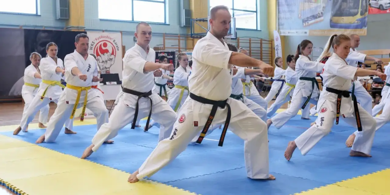 Grupa osób w kimonach ćwiczy karate na niebiesko-żółtej macie w sali gimnastycznej.