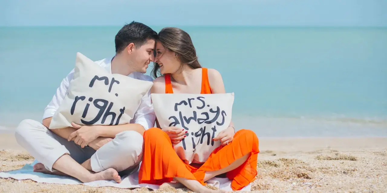 Para zakochanych na plaży z poduszkami "Mr. Right" i "Mrs. Always Right". Ich uśmiechy i bliskość symbolizują idealny **kompromis w związku**.
