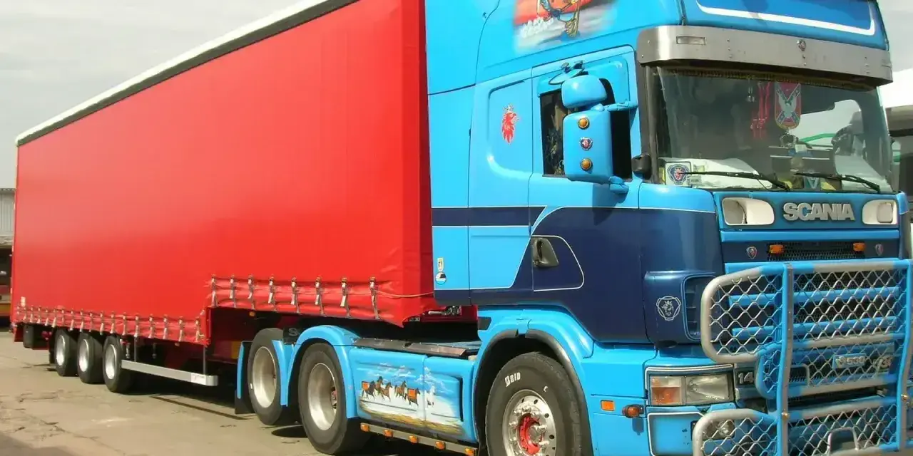 Naczepa jumbo w jaskrawym kolorze czerwonym, ciągnięta przez niebieską Scanię z malowidłami.