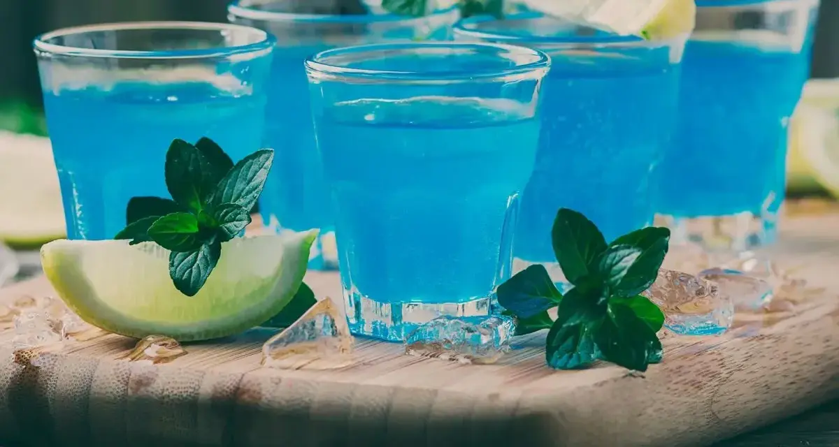 Kilka shotów z drinkiem blue curacao, ozdobionych limonką i miętą, na drewnianej desce z lodem.