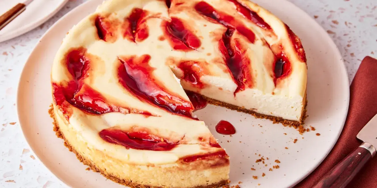 Ein saftiger american cheesecake mit keksboden, verziert mit roten Fruchtwirbeln. Ein Stück fehlt bereits.
