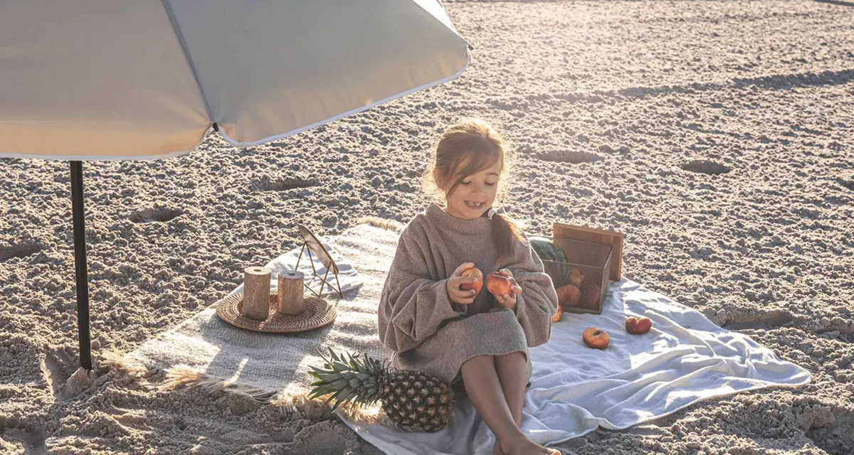 Dziewczynka na plaży pod parasolem, jedząca owoce. Idealny zestaw na wakacje nad morze z dzieckiem: koc, owoce, ananas.
