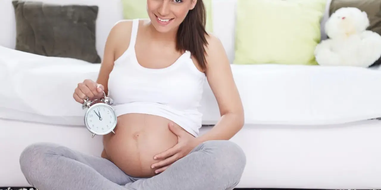 Uśmiechnięta kobieta w 40 tygodniu ciąży trzyma budzik. Czas na poród!