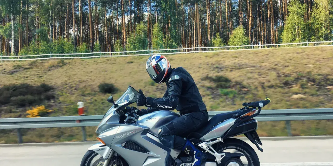 Motocyklista w kasku jedzie na srebrnym Honda VFR 800 wzdłuż leśnej drogi.