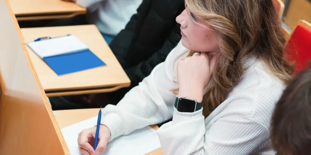 Studentka pisze notatki na wykładzie, opierając podbródek na dłoni.