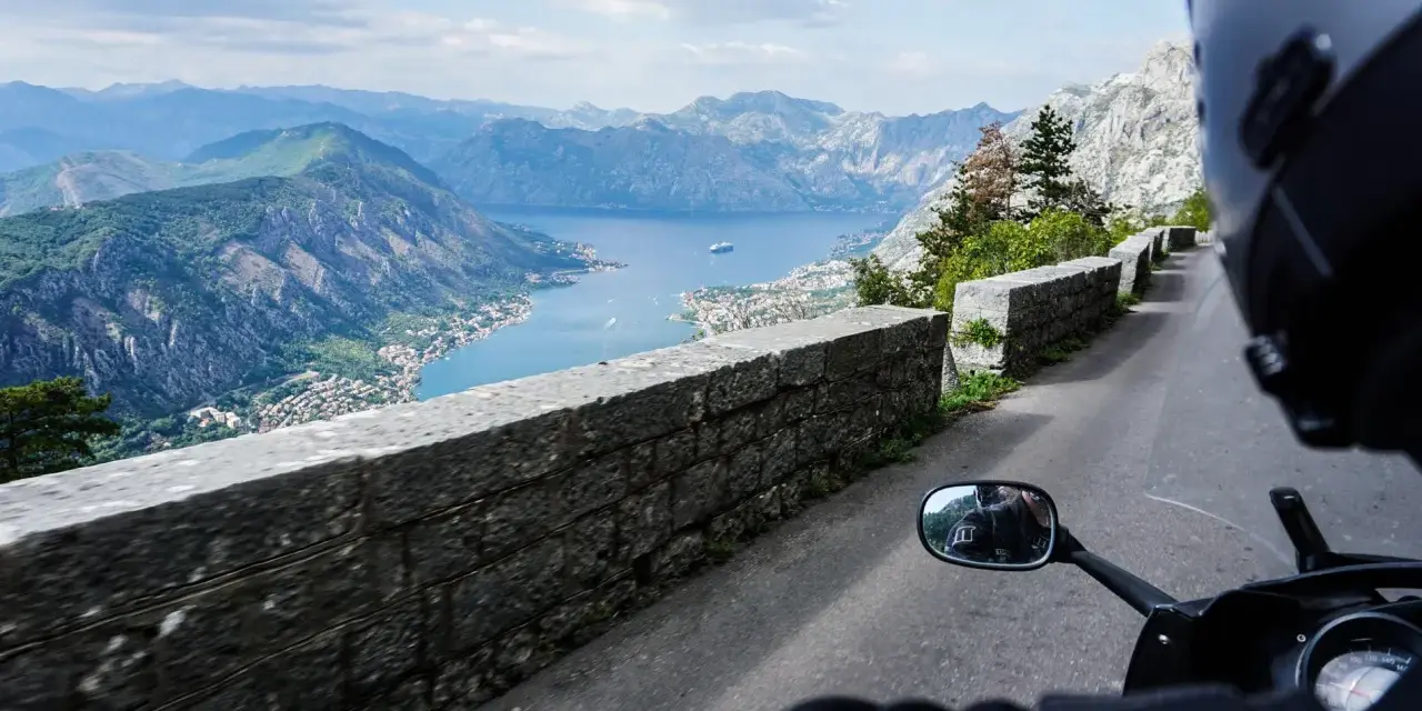 Widok z motocykla na Zatokę Kotorską w Czarnogórze. Kręta droga z kamiennym murem i zapierająca dech w piersiach panorama gór i morza.