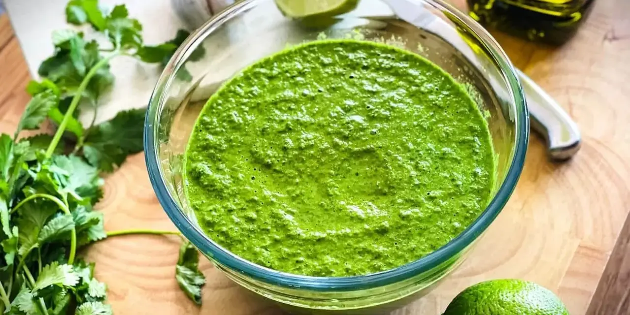 Frisch zubereitetes mojo verde in einer Glasschüssel, umgeben von Koriander, Knoblauch, Limetten und Olivenöl.