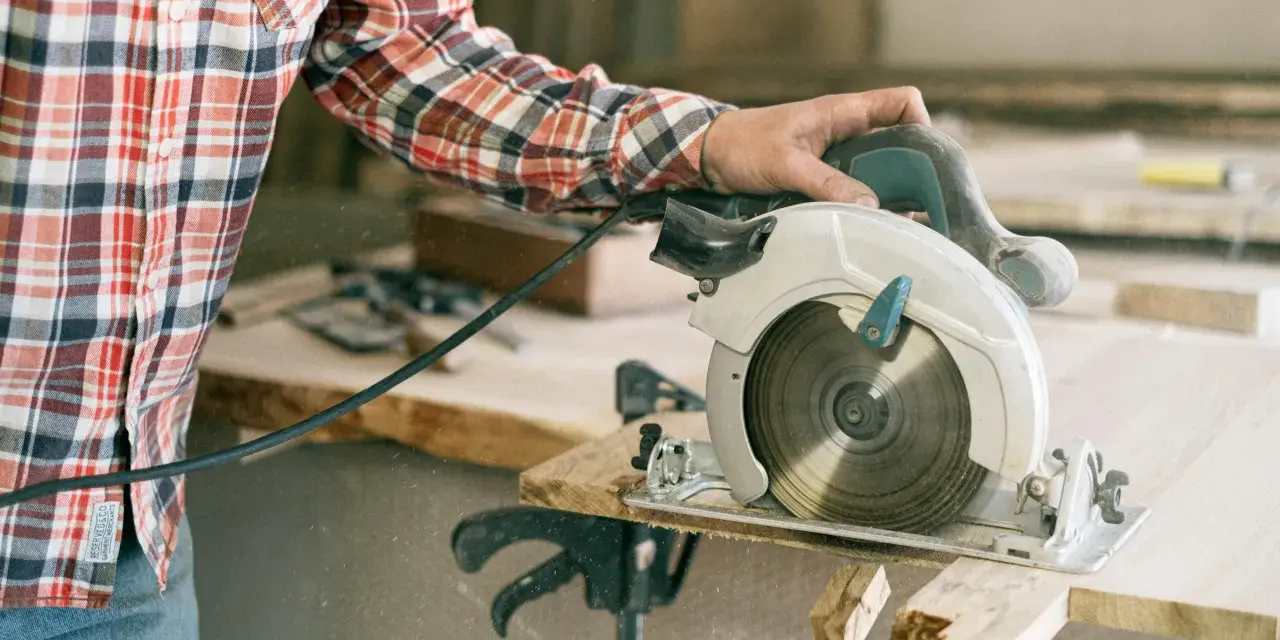 A person in a plaid shirt demonstrates how to use a circular saw, cutting wood with sawdust flying.