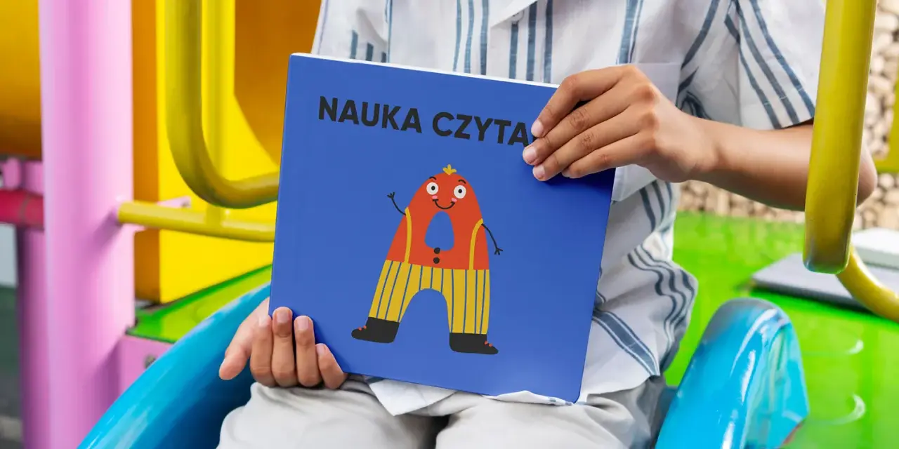 Chłopiec trzyma książkę "Nauka czytania dla 6 latka" z zabawną literką "A" na okładce.