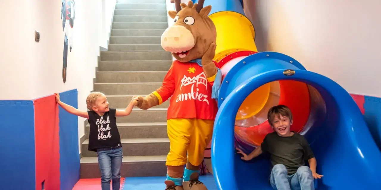 Mascot and two children in a colorful indoor playground with a slide and stairs.