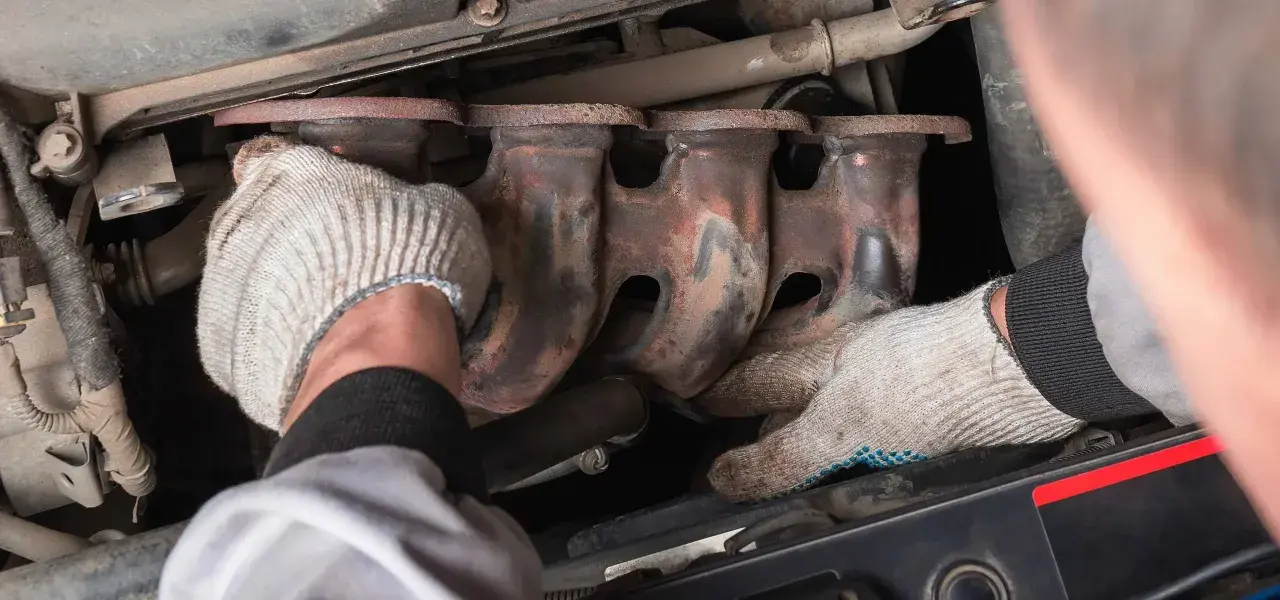 Mechanic's gloved hands work on a rusty exhaust manifold, possibly to fix an engine ticking noise.