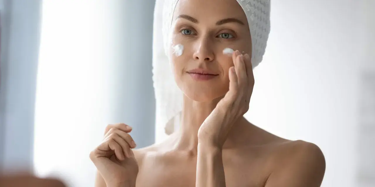 Femme appliquant de la crème sur son visage pour lutter contre la xerose cutanée. Elle a une serviette sur la tête.