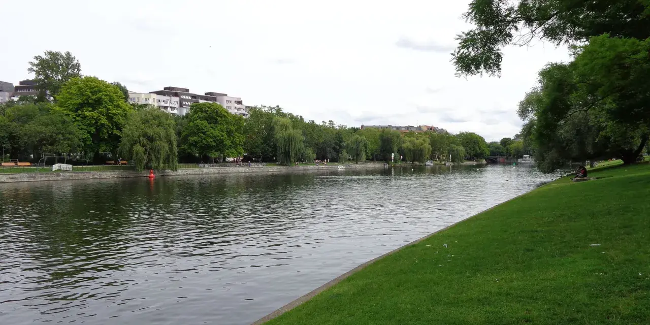 Grüne Ufer am Landwehrkanal Berlin mit Bäumen, Gebäuden und Menschen, die sich auf der Wiese entspannen.