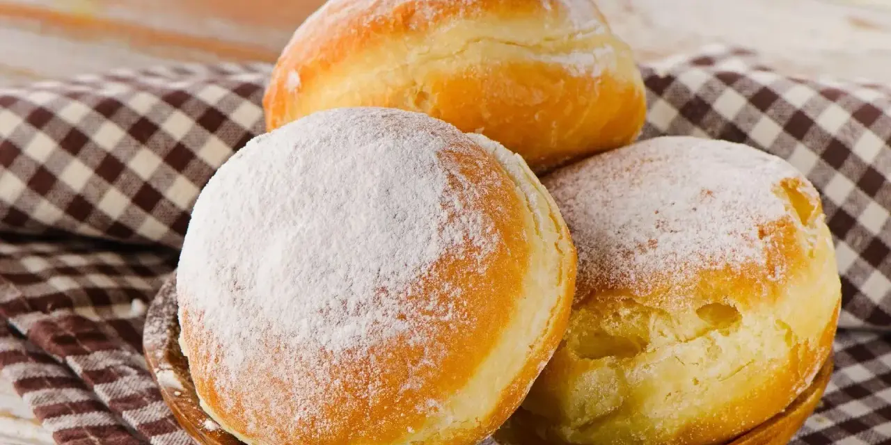 Trzy puszyste pączki, dwa posypane cukrem pudrem, leżą na drewnianej misce. Idealne na tłusty czwartek, te fit pączki kuszą apetycznym wyglądem.