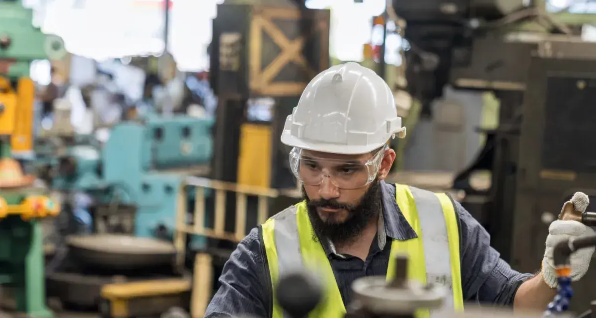 Obrero con casco y gafas de seguridad trabajando en una máquina en una fábrica, parte esencial de la manufactura moderna.