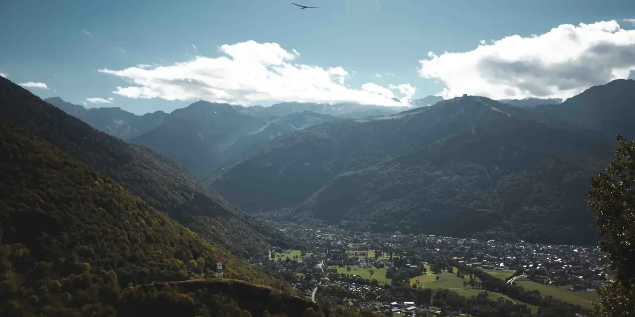 Vue aérienne d'une vallée verdoyante avec un village niché, entouré de montagnes majestueuses. Un planeur plane dans le ciel bleu, rappelant les paysages de Toulouse et ses environs.