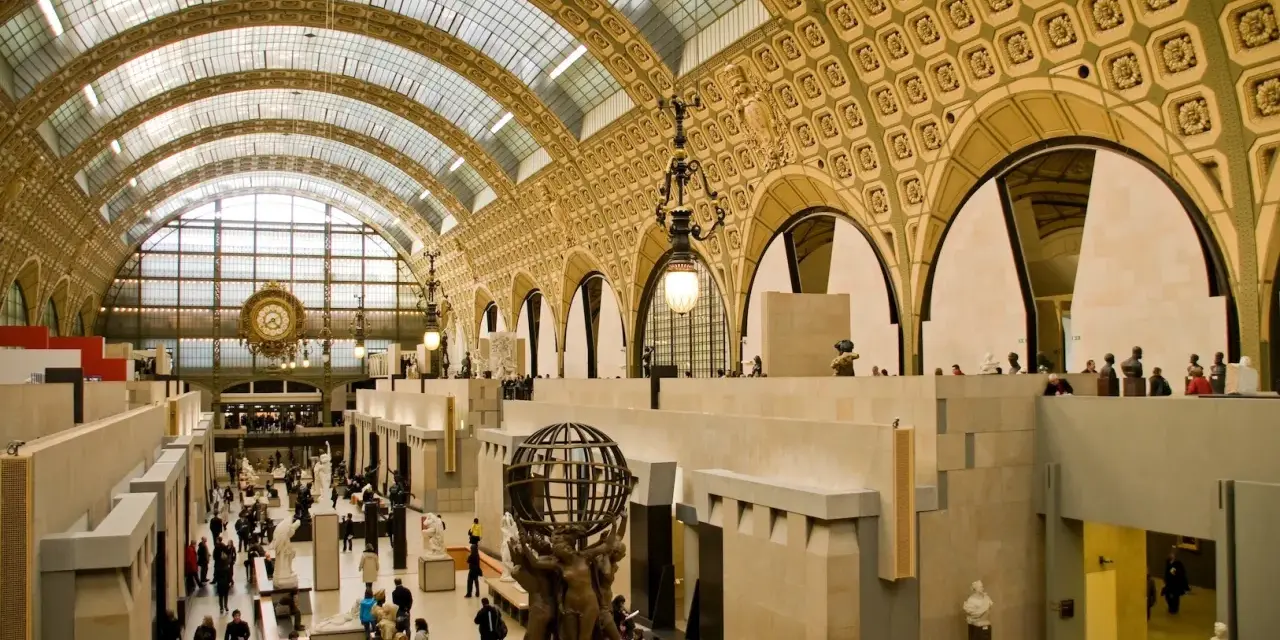 Interior del museo d'Orsay, con su impresionante techo de cristal y arcos dorados. Visitantes pasean entre esculturas y obras de arte.