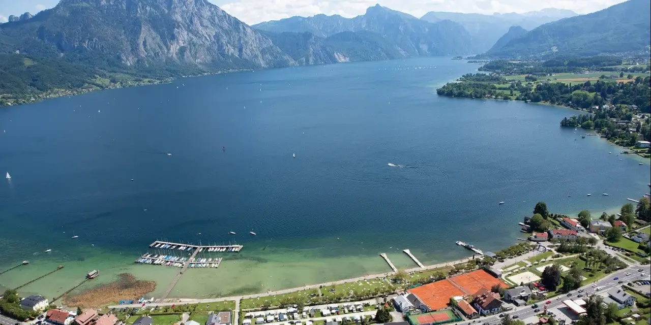 Idyllischer **Camping Traunsee** mit Blick auf den See und die Berge. Boote liegen im Hafen, Tennisplätze und Wohnwagen sind zu sehen.