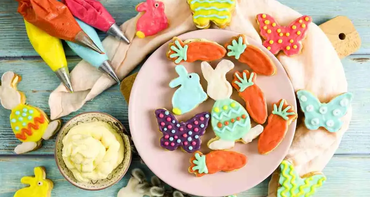 Bunte Osterkekse in Hasen-, Karotten- und Schmetterlingsform, bereit für ein einfaches Osterkekse Rezept. Spritzbeutel mit Zuckerguss liegen bereit.