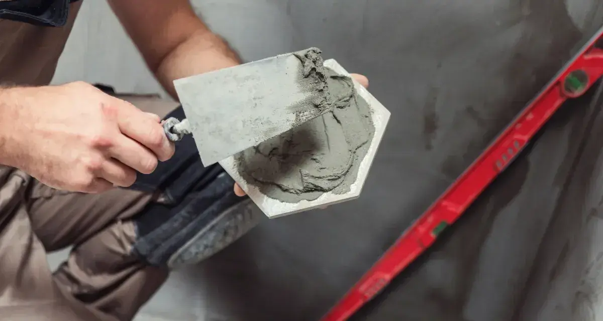 Majster nakłada klej na płytkę, przygotowując ją do ułożenia. Obok leży poziomica, a w tle widać materiał, który może być matą pod płytki na OSB.