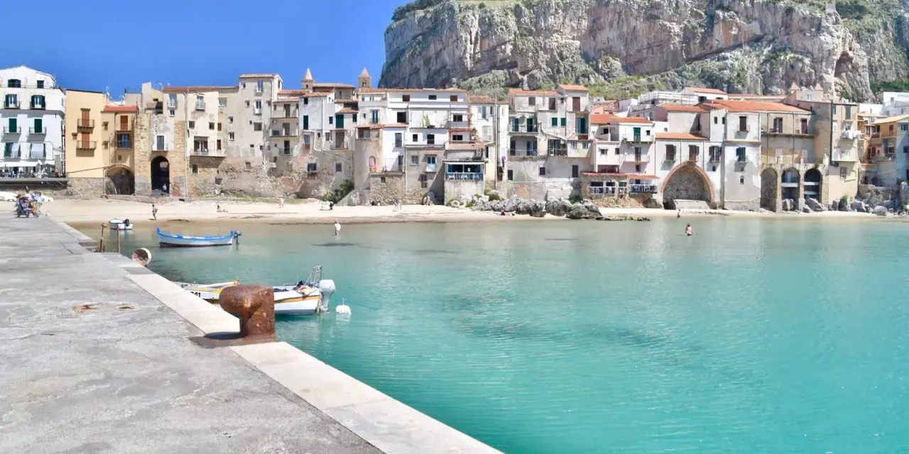 Coastal town with buildings nestled against a cliff, turquoise water, and boats. Perfect for a 10 days in Sicily adventure.