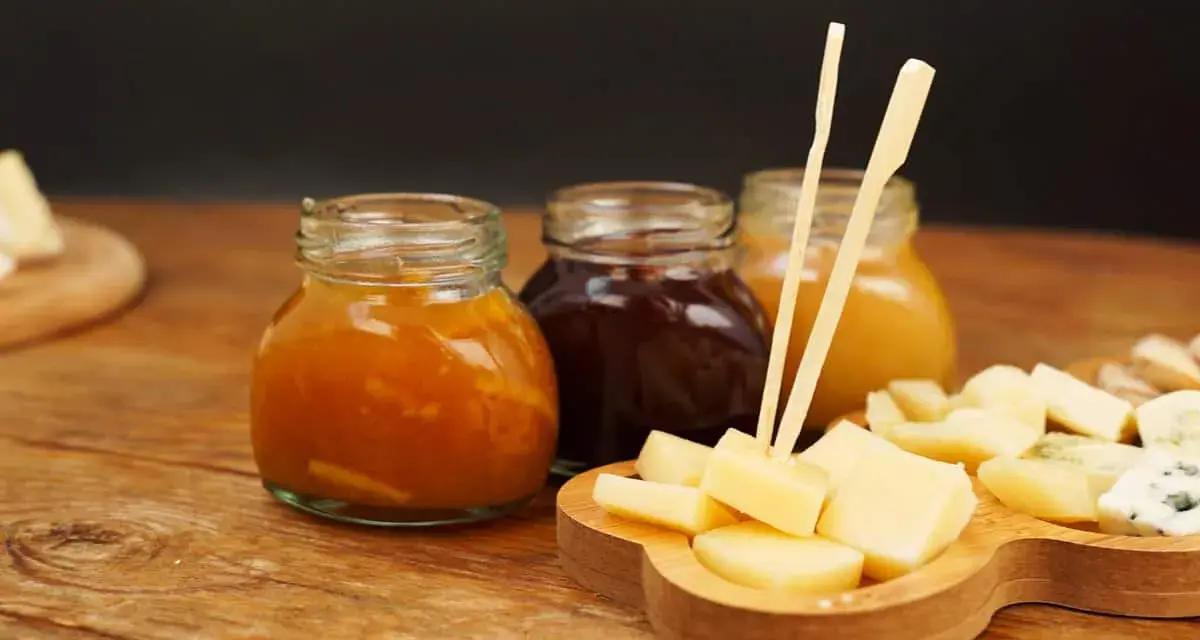 Tre vasetti di marmellata, uno arancione, uno scuro e uno giallo, accompagnano un tagliere di formaggio a cubetti, pronto per essere gustato.