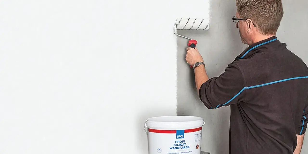 Mann streicht mit einer Farbrolle eine Wand weiß. Der Eimer mit der "Profi Silikat Wandfarbe" steht daneben. Perfekt für das Streichen bei Auszug.