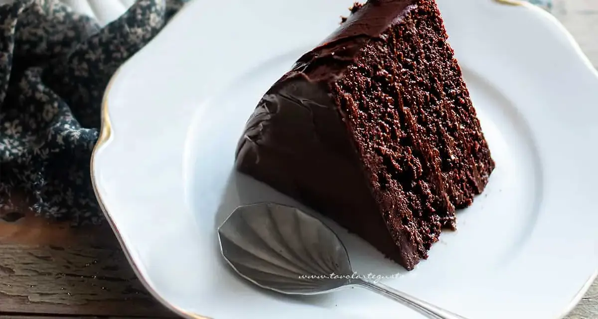 Una fetta di torta al cioccolato morbida e umida, ricoperta di glassa lucida, su un piatto bianco con un cucchiaino.
