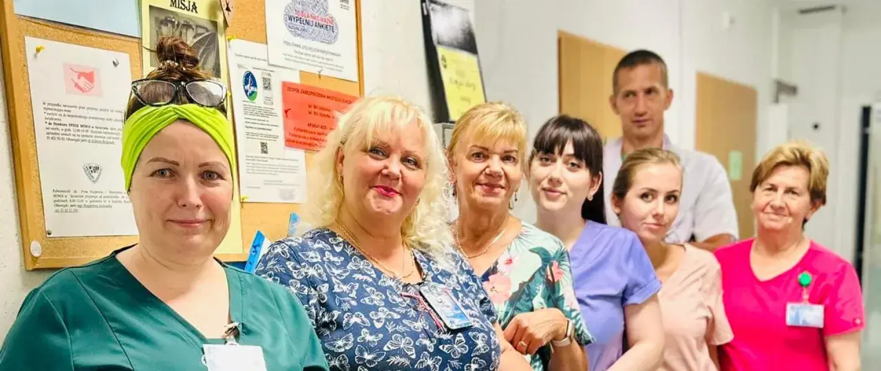 Grupa uśmiechniętych pracowników medycznych w szpitalu, na tle tablicy ogłoszeń.