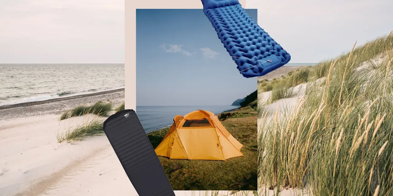Zwei Isomatten, eine blaue aufblasbare und eine schwarze Schaumstoffmatte, liegen neben einem orangefarbenen Zelt am Strand.
