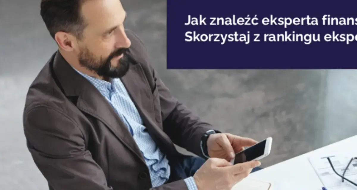 Mężczyzna z brodą trzyma smartfon, siedząc przy biurku z dokumentami.