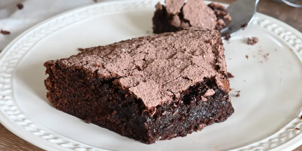 Una fetta di torta al cioccolato fondente, cosparsa di cacao, su un piatto bianco. Un pezzetto è stato staccato con una forchetta.