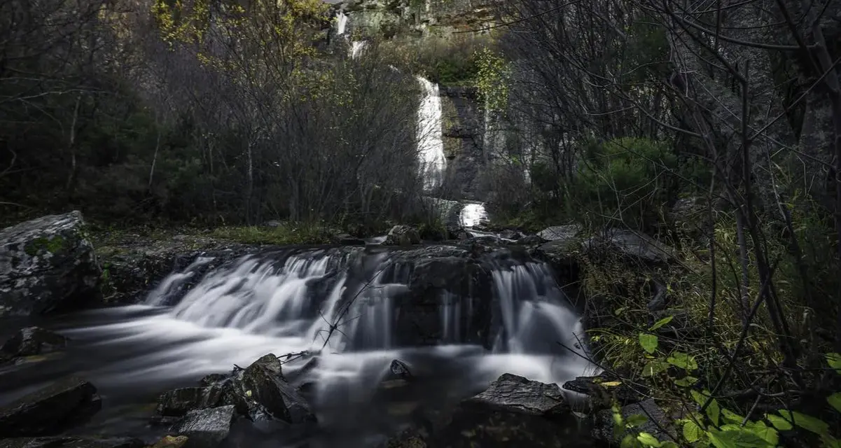 Cascada en el **río Bornova**, con aguas sedosas cayendo entre rocas y vegetación otoñal, bajo un cielo nublado.