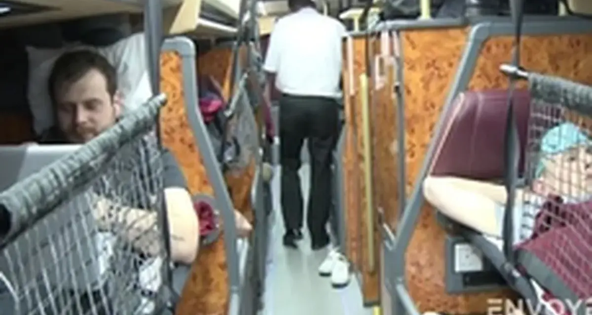 Intérieur d'un bus avec des couchettes. Un homme en blanc marche dans l'allée, tandis que des passagers se reposent dans leurs couchettes.