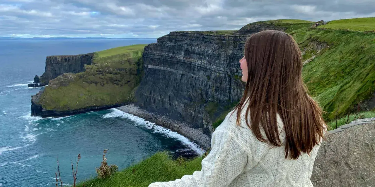 A woman in a cream cable-knit sweater gazes at the dramatic Cliffs of Moher, a breathtaking highlight of many tours of Ireland.