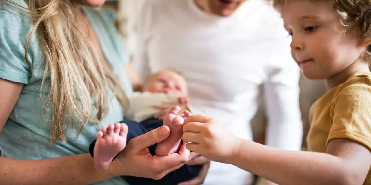 Ein älteres Kind berührt neugierig die Füßchen des Babys. Ein Hauch von eifersucht unter geschwistern liegt in der Luft, während die Eltern das neue Familienmitglied halten.