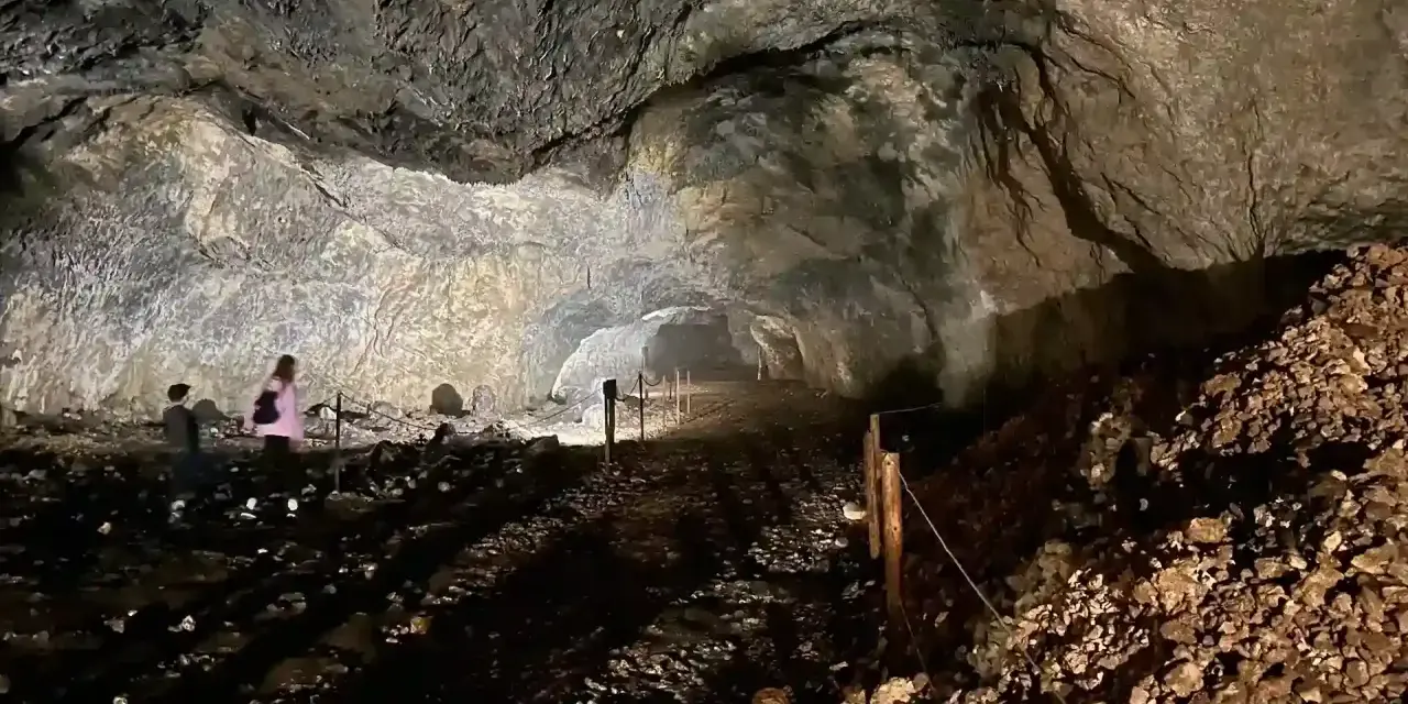 Wnętrze jaskini ciemnej, z kamienistym dnem i skalnymi ścianami. W oddali widać dwie osoby zwiedzające.