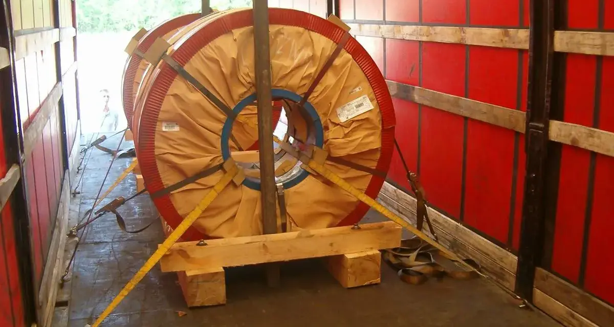 Dwa wielkie zwoje metalu zabezpieczone drewnianym mocowaniem blokowym i pasami transportowymi w naczepie.