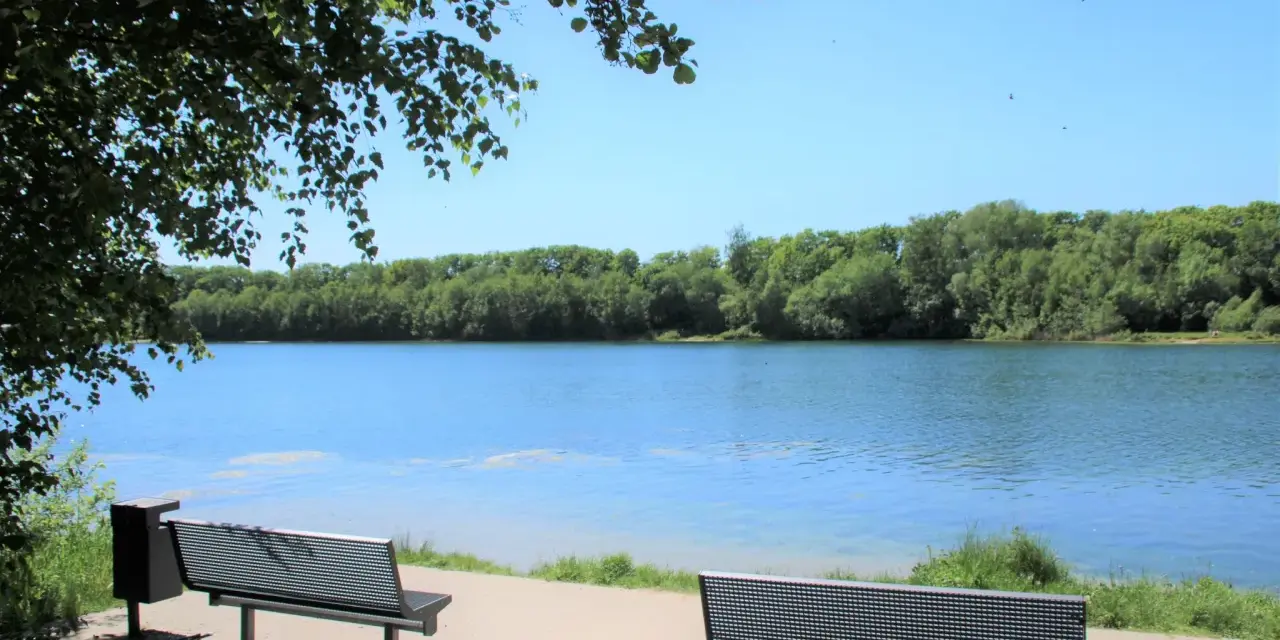 Zwei Bänke laden zum Verweilen am Ufer des halener Badesees ein. Bäume säumen das klare Wasser unter blauem Himmel.