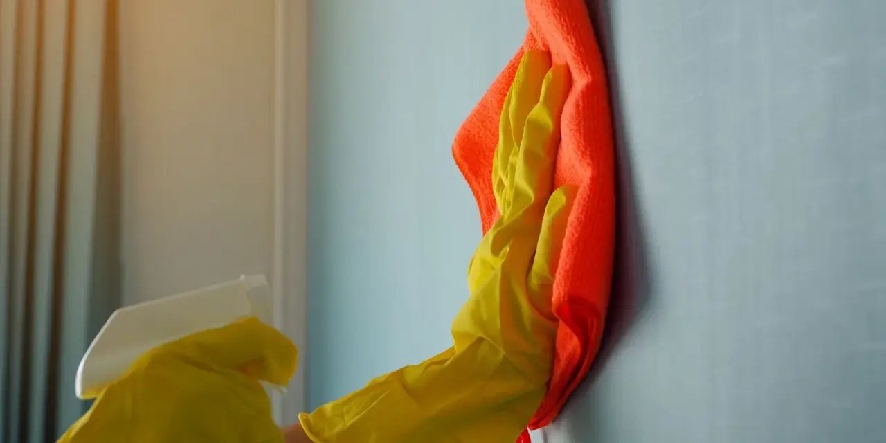 Ręka w żółtej rękawiczce z pomarańczową ściereczką czyści ścianę. Dowiedz się, czym zmyć farbę ceramiczną, by przywrócić jej blask.