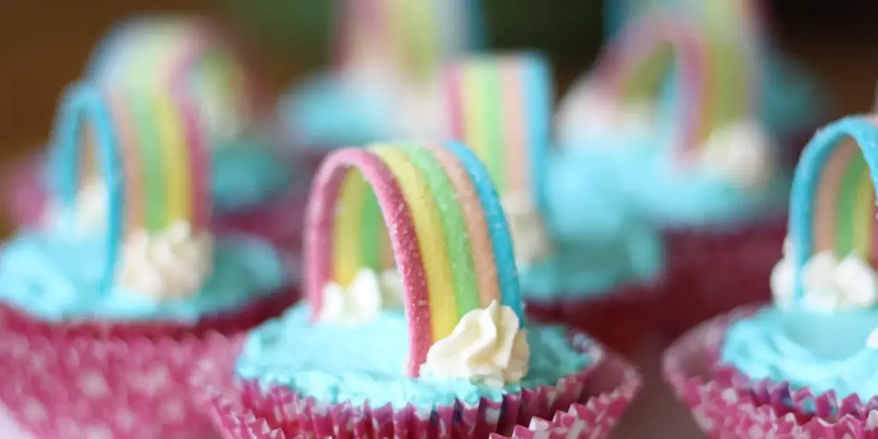 Sju cupcakes med ljusblå regnbågs frosting och godisregnbågar. Rosa muffinsformar med mönster.