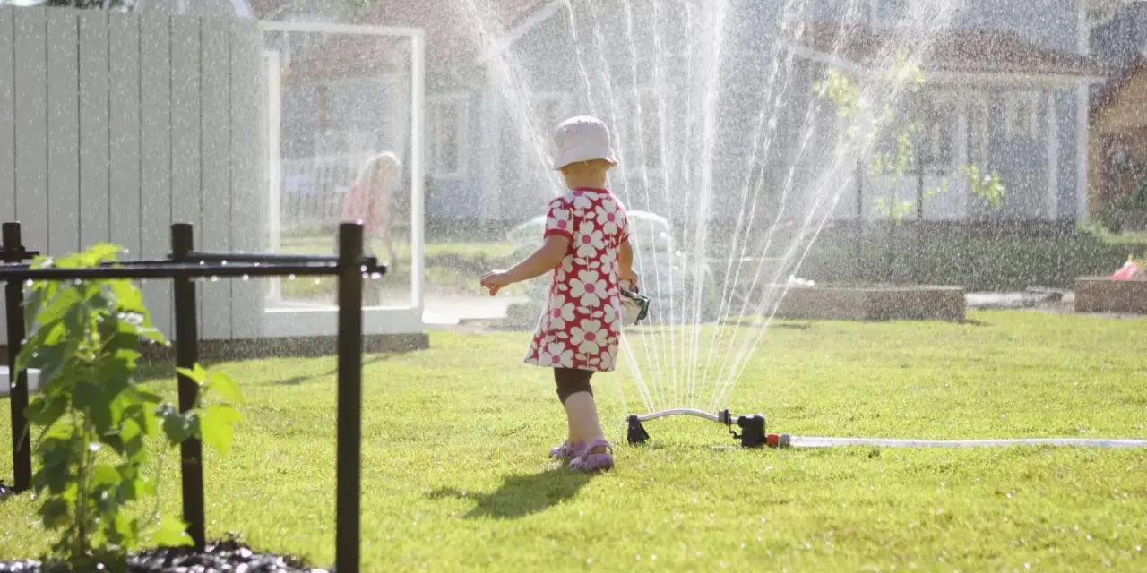 Lekfull **tipspromenad barn** i sommarregn från en vattenspridare. Ett litet barn i blommig klänning njuter av vattnet på gräsmattan.