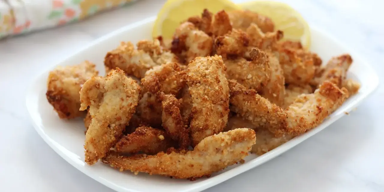Straccetti di pollo in friggitrice ad aria, dorati e croccanti, serviti su un piatto bianco con spicchi di limone.