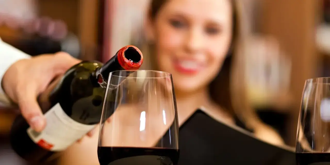Kelner nalewa wino, a uśmiechnięta kobieta czeka na kieliszek. Wino zawiera siarczyny, które pomagają w jego konserwacji.